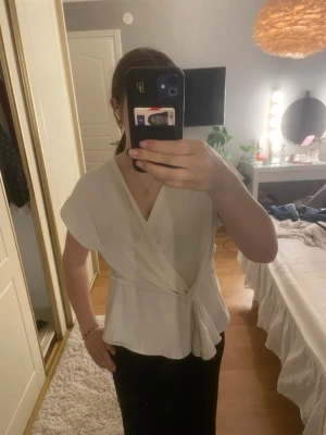 Vit omlottopp från Zara - Säljer ett av mina favoritplagg då den tyvärr inte passar längre💞Den är lite liten i storlek men inte jätte liten, sitter bra på någon nånstans mellan storlek L och M men går även hur bra som helst för de som har M eller liknande bara att den sitter lite lösare💘En stilren vit omlottopp från Zara med knyt i midjan och v-ringad hals. Toppen har korta ärmar och ett mjukt, lätt material som ger ett snyggt fall. Perfekt att styla med både jeans och kjol för en trendig look.