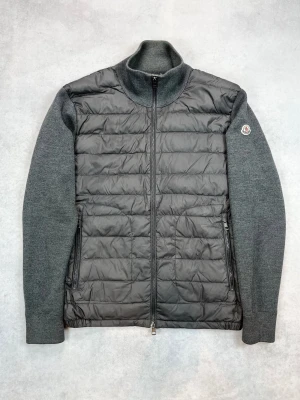 Grå cardiganjacka från Moncler - Snygg grå cardiganjacka från Moncler med quiltad front i nylon och stickade ärmar samt rygg i ullmix. Jackan har hög krage, hel dragkedja och två sidofickor med dragkedja. Moncler-logga på vänster ärm för extra stilpoäng.