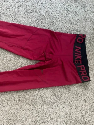 Vinröda Nike PRO Dri-FIT träningstights - Säljer ett par vinröda Nike Dri-FIT träningstights i storlek M. Tightsen har hög midja med bred resår och är tillverkade i ett stretchigt funktionsmaterial som transporterar bort fukt. Perfekta för gymmet eller löpning.