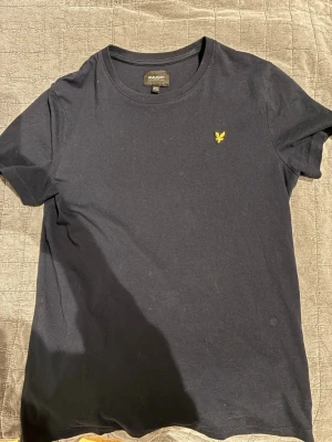 Svart t-shirt från Lyle & Scott - En stilren svart t-shirt från Lyle & Scott med klassisk rund hals och korta ärmar. På bröstet sitter den ikoniska gula örnloggan broderad. Tillverkad i mjuk bomull för en skön känsla och enkel att matcha med allt. OBS FINNS EN DEFEKT I HÖGRA ARMHÅLAN(litet hål)