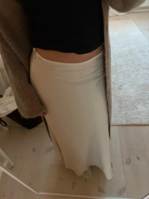 Beige långkjol i satin - Säljer en beige långkjol i mjukt satinliknande material. Kjolen har en enkel och stilren design med lätt utsvängd form som ger en snygg siluett. Perfekt att styla med både stickade tröjor och crop tops.