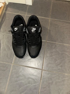 Nike Air Max 90  - Svarta Nike Air Max 90 sneakers med vit sula och vit swoosh. Ovandelen är i mesh och syntetmaterial för en sportig look. Klassisk Air Max-dämpning i sulan och tydlig logga på sidan. Perfekta för dig som gillar streetwear och sportig stil.