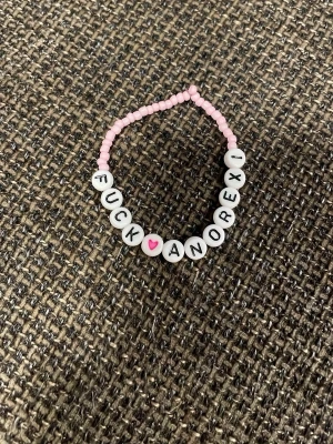 Pärlarmband med budskap och hjärta - Ett handgjort armband med små rosa pärlor och vita bokstavspärlor som bildar texten 'FUCK ANOREXI' samt ett rosa hjärta. Armbandet har en stretchig tråd och är perfekt för dig som vill bära ett statement.