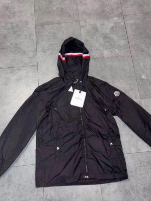 Svart vindjacka från Moncler - Svart vindjacka från Moncler med huva och dragkedja framtill. Jackan har röda, vita och blå detaljer på huvan samt Moncler-logga på ärmen. Tillverkad i lätt och glansigt material, perfekt för blåsiga dagar.