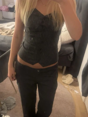 Grå randig korsettopp - Säljer en grå korsettopp med tunna vita ränder och knappar framtill. Toppen har en v-ringad skärning och är figurnära med en croppad passform. Perfekt att styla med både jeans och kostymbyxor för en trendig look.