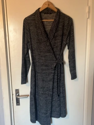 Svartvit omlottklänning med knyt - Mönstrad svartvit omlottklänning med knyt i midjan och långa ärmar. Klänningen har ett diskret randigt mönster och är i mjukt, stretchigt material. Perfekt för dig som gillar en enkel men snygg look.