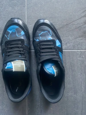 Valentino sneakers med blått mönster - Säljer ett par snygga sneakers från Valentino med svart och blått kamouflagemönster. Skorna har svarta skosnören och en låg profil. Materialet är en mix av läder och textil, perfekt för dig som vill sticka ut med ett exklusivt märke. Går att diskutera priset!!