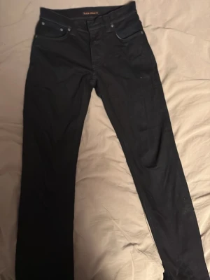 Svarta jeans från Nudie Jeans - Säljer ett par svarta jeans från Nudie Jeans med klassisk femficksmodell och raka ben. Jeansen har svarta sömmar och subtil logga på bakfickan. Materialet är mjukt, vilket gör dem riktigt sköna att bära. Perfekta för en clean och stilren look.