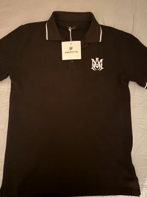 Svart pikétröja från Amiri - Snygg svart pikétröja från Amiri med vit logga på bröstet och vita detaljer på kragen. Klassisk pikéknäppning framtill och korta ärmar. Perfekt för en clean och stilren look. Pris kan diskutera 