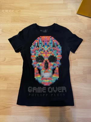 Svart t-shirt med döskalle från Philipp Plein - Limiterad svart t-shirt från Philipp Plein med en stor färgglad döskalle i pixelstil framtill, dekorerad med glittriga paljetter. Texten 'GAME OVER' och varumärket syns under motivet. Perfekt för dig som gillar statement-plagg och unika detaljer.