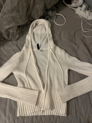 Vit ribbstickad hoodie från Divided - Säljer en vit ribbstickad hoodie från Divided med dragkedja framtill. Tröjan har långa ärmar, åtsittande passform och en stor huva. Perfekt för lager på lager och enkel att matcha med olika outfits.