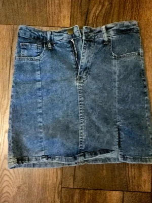 Blå jeanskjol med dragkedja - Säljer en klassisk blå jeanskjol i kort modell med dragkedja och knapp framtill. Kjolen har fickor både fram och bak samt bälteshällor. Perfekt till vardags för en avslappnad och trendig look.