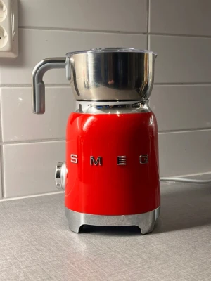 Röd mjölkskummare från Smeg - Säljer en snygg röd mjölkskummare från Smeg i retrostil. Maskinen har en blank metallkanna med handtag och genomskinligt lock. Perfekt för att fixa cappuccino eller varm choklad hemma. Står stadigt på bänken och har en klassisk Smeg-design.