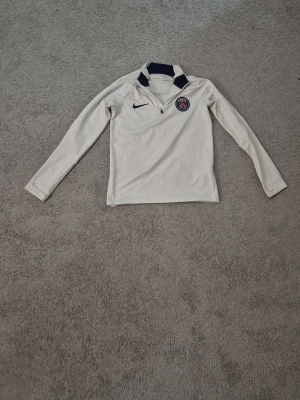 Vit PSG x Nike tröja - Vit långärmad tröja från Nike med PSG-logga på bröstet och marinblå detaljer vid kragen. Tröjan har half zip-dragkedja och är tillverkad i ett sportigt syntetmaterial som känns lätt och bekvämt. Perfekt för dig som gillar sportig stil.