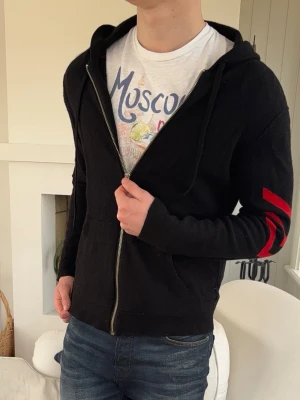 Zadig Voltaire Kaschmir hoodie zip up - Snygg svart hoodie med dragkedja och huva. På vänster ärm finns tre röda ränder som ger en cool kontrast. Hoodien har fickor framtill och är tillverkad i ett mjukt material som känns skönt mot huden. Perfekt för en avslappnad streetwear-look.