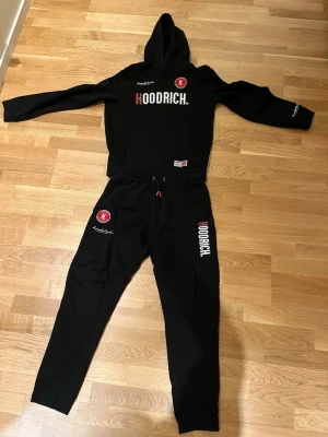 Svarta mjukisbyxor från Hoodrich(xs) - Säljer ett par svarta mjukisbyxor från Hoodrich med vit och röd logga på benet och tryckt text längs sidan. Byxorna har dragsko i midjan och muddar vid bensluten. Perfekta för en chill och street-inspirerad look.