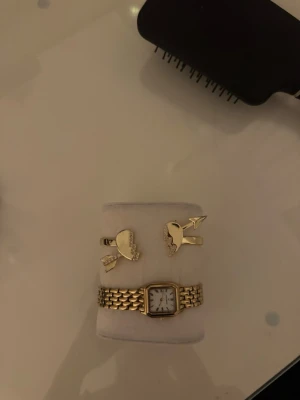 Guldigt armband med hjärtmotiv - Säljer ett coolt armband i guldfärgad metall med två hjärtformade detaljer och pilmotiv. Armbandet har en öppen design och är perfekt för dig som gillar unika accessoarer med lite edge. Passar dig som vill sticka ut och addera något extra till din stil.