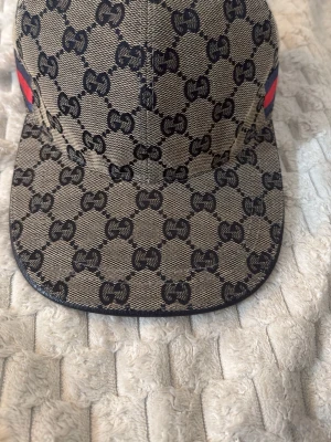 Beige keps med GG-mönster från Gucci - Snygg keps från Gucci i beige canvas med klassiskt GG-monogram över hela kepsen. Har en böjd skärm och ikoniska röd-blå ränder på sidan. Märkeslapp medföljer.Storlek M=58 cm.Perfekt för dig som vill sticka ut med lyxig streetstyle.