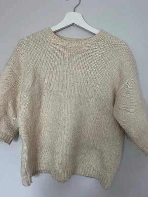 Beige stickad tröja med rund hals - Mysig beige stickad tröja med rund halsringning och ribbade muddar vid ärmslut och nederkant. Tröjan har en avslappnad passform och är tillverkad i ett mjukt material som passar perfekt för kyligare dagar.