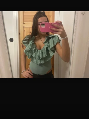 Grön volang topp med djup v-ringning - Säljer en snygg grön topp med volangdetaljer och djup v-ringning. Toppen är tight och har ärmlös design, perfekt för att skapa en cool och trendig look. Materialet känns som bomull och polyester, vilket gör den bekväm att bära.