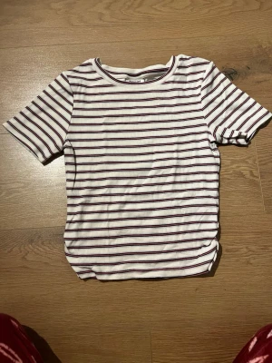 Randig t-shirt från Kappahl - Säljer en vit t-shirt från Kappahl med tunna horisontella ränder i rött, svart och rosa. T-shirten har rund halsringning, korta ärmar och är gjord i mjuk bomull med ribbad struktur. Perfekt till en avslappnad vardagsstil.