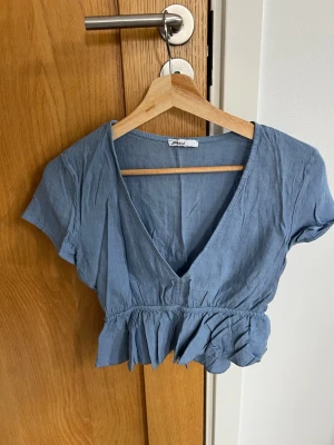 Blå croppad topp i linne, från Subdued - Snygg blå croppad topp, från Subdued med v-ringning och korta ärmar. Toppen har volangdetalj i midjan och är tillverkad i ett mjukt linnetyg. Perfekt för dig som gillar en avslappnad men trendig stil.