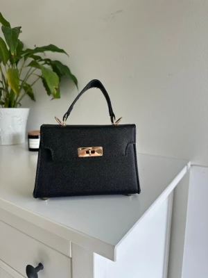 Svart minihandväska med gulddetaljer - Stilren svart minihandväska i läderimitation med guldfärgat vridlås och handtag. Väskan har en klassisk, rektangulär form och små guldfärgade fötter undertill. Perfekt accessoar för att lyfta din outfit.