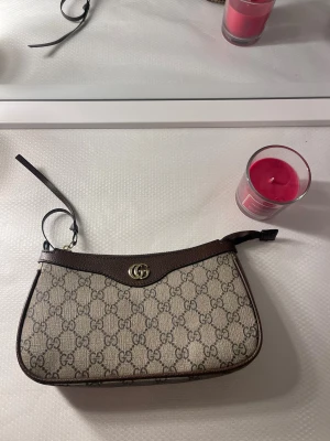  beige GG canvas axelremsväska - Säljer en snygg beige axelremsväska med klassiskt GG-monogram i canvas och mörkbruna läderdetaljer. Väskan har guldfärgad logga framtill och en smal axelrem i läder. Perfekt för dig som gillar lyxiga accessoarer med ikonisk design.