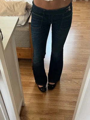 Mörkblå jeans med sidoknappar - Säljer ett par mörkblå bootcut jeans som är lågmidjade med dubbla knappar på sidan. Fickorna är vanliga och det finns inga tecken på användning💓💓