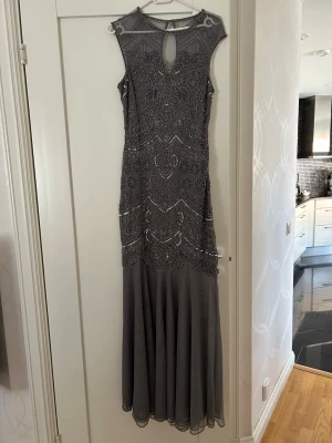 Grå långklänning med pärlor - Elegant grå långklänning med vackra pärlbroderier och detaljerad spets upptill. Klänningen är ärmlös och har en lätt transparent nederdel i chiffongliknande material. Perfekt för speciella tillfällen och ger en lyxig känsla.