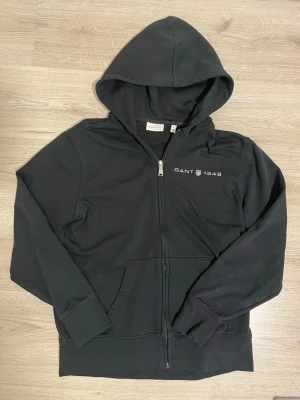 Svart hoodie med dragkedja från Gant - Svart hoodie från Gant med dragkedja framtill och broderad logga 'GANT 1949' på bröstet. Klassisk huva och känguruficka. Mjuk bomullskänsla och ribbade muddar vid ärmslut och nederkant. Perfekt för en chill och stilren look. Bredd 49 cm, längd 65 cm