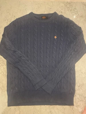 Blå kabelstickad tröja Morris - Kabelstickad blå tröja från Morris i 100% pima bomull. Klassisk rund halsringning, ribbade muddar och en liten orange logga på bröstet. Perfekt för dig som gillar stilrena och tidlösa plagg.