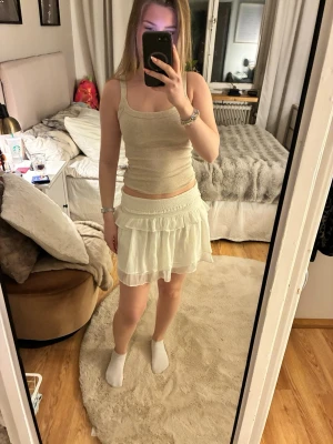 Vit volangkjol med ribbad midja - Säljer en kort vit kjol med volang och ribbad midja. Kjolen har en lätt och luftig känsla, perfekt för varma dagar. Den är gjord i ett tunt material som ger ett snyggt fall och en feminin look.