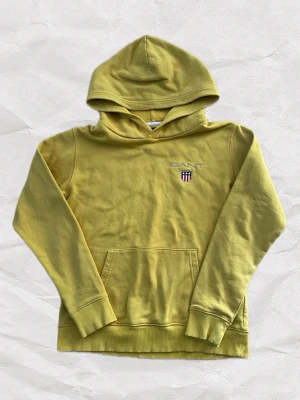 Gul Gant Hoodie - Snygg gul hoodie från GANT med broderad logga och sköld på bröstet. Ribbade muddar vid ärmar och nederkant. Tillverkad i mjuk bomullsblandning.