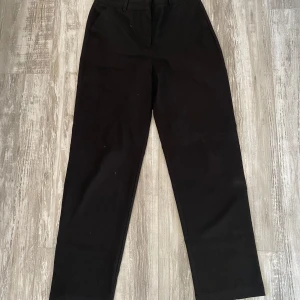 Svarta raka kostymbyxor - Stilrena svarta kostymbyxor med rak passform och hällor för skärp. Klassisk design som funkar till många olika looks. Byxorna har diskreta fickor och stängs med dragkedja och knapp framtill. Perfekta för dig som gillar en clean och enkel stil.