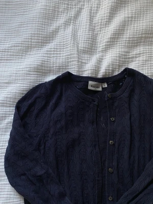 Mörkblå kofta från Kappahl - Snygg mörkblå kofta från Kappahl med rund halsringning och stickat mönster. Koftan har knappar framtill och ribbade muddar vid ärmslut och nederkant. Perfekt att slänga över en topp för en chill look. 