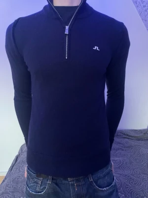 Mörkblå stickad tröja från J.Lindeberg - Snygg mörkblå stickad tröja från J.Lindeberg med halv dragkedja och diskret logga på bröstet. Tröjan har lång ärm, ribbade muddar och en smal passform. Perfekt till jeans för en clean look.