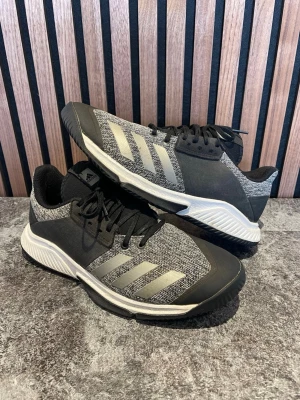 Svarta/grå Adidas sneakers - Snygga sneakers från Adidas i svart med grå textil partier. Skorna har svart snörning och en svart sula med vita detaljer. Ovandelen är i textil och syntetmaterial. Hälen i skon är sliten annars i bra skick