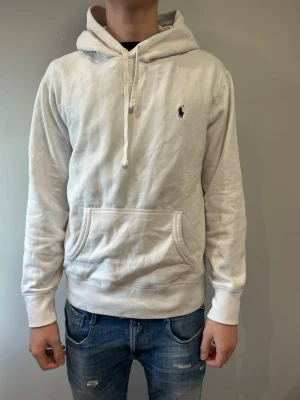 Vit hoodie från Polo Ralph Lauren - Säljer en stilren vit hoodie från Polo Ralph Lauren med klassisk huva och dragsko. Tröjan har en känguruficka framtill och den ikoniska lilla loggan broderad på bröstet. Perfekt till jeans för en avslappnad look.