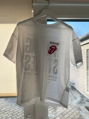 Rolling Stones t-shirt från H&M - Vit t-shirt med Rolling Stones-tryck fram och stort konsertmotiv på ryggen. Klassisk rund hals och korta ärmar. Ikonisk röd tunga-logga på bröstet. Perfekt för dig som gillar bandmerch och vill sticka ut med en cool och tidlös stil.