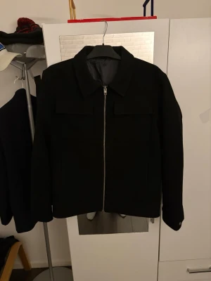 Svart overshirt med dragkedja - Stilren svart overshirt med klassisk krage och silverfärgad dragkedja framtill. Två bröstfickor med lock ger en clean look. Overshirten har långa ärmar med knapp vid ärmslut och är tillverkad i ett tjockare tyg som känns robust.