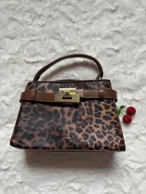 Leopardmönstrad handväska med gulddetalj - Snygg handväska i leopardmönstrat konstläder med bruna detaljer och guldfärgat spänne. Väskan har en kort bärrem och en stilren, boxig form som ger en trendig look. Perfekt accessoar för att lyfta din outfit.