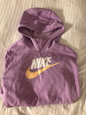 Lila Nike hoodie med logga - Säljer en helt oanvänd lila hoodie från Nike med stor vit och gul logga på bröstet. Hoodien har huva, känguruficka och långa ärmar. I ungdom/ barn storlek 156-166/XL