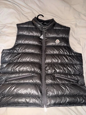 Mörkgrå  dunväst från Moncler - Snygg mörkgrå dunväst från Moncler med klassisk quiltad design och dragkedja framtill. Västen har två snedställda fickor med dragkedja och Moncler-logga på bröstet. Perfekt för lager-på-lager och håller dig varm med stil.