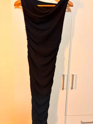 Svart glittrig långklänning - Säljer en svart långklänning med glittrigt tyg och draperad design. Klänningen har en elegant form och är perfekt för dig som vill sticka ut. Materialet är syntet och klänningen har en slits som ger extra attityd. Passar dig som gillar glam och stil.