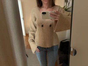 Beige omlottkofta med vida ärmar - Trendig beige omlottkofta med två dekorativa knappar framtill och extra vida ärmar. Koftan har en djup v-ringning och är tillverkad i ett mjukt stickat material. Perfekt för en avslappnad och stilren look.