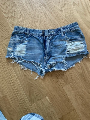 Blå slitna jeansshorts Levi's - Säljer ett par blå jeansshorts från Levi's med slitna detaljer och fransig kant. Klassisk femficksmodell med låg midja och knappgylf. Perfekta för sommaren och ger en cool, avslappnad look.