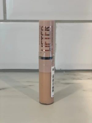Maybelline Lifter Concealer 20 - Maybelline Lifter Concealer i nyansen 20. Kommer i en smidig tub med skruvlock och applikator. Färgen är ljusbeige och förpackningen är i plast med rosatonad etikett och metallisk text. Perfekt för att täcka mörka ringar och ojämnheter.
