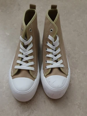 Sneakers - Snygga höga sneakers i beige canvas med vita gummisulor och tåhättor. Skorna har vita skosnören och ljusgröna detaljer runt öppningen. Klassisk och stilren design som passar till många outfits. Finns några fläckar.