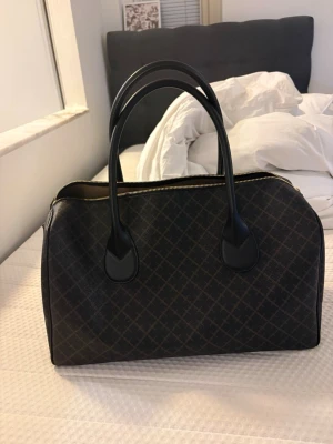 Malene Birger väska  - Snygg mörkbrun handväska med diskret rutmönster och svarta handtag. Väskan har guldfärgade detaljer och dragkedja upptill. Klassisk, avlång form som rymmer mycket. Materialet ser ut att vara konstläder eller canvas. Nypris 4900kr, mitt pris 3500kr!